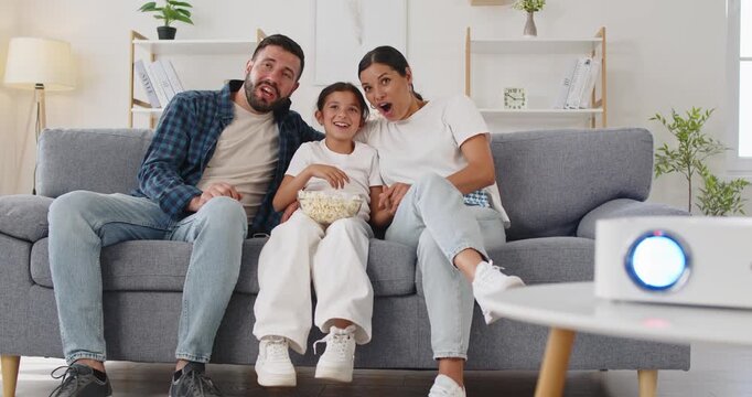 Family watching movie at home with projector. Parents and child sit on a cozy sofa couch in the living room, reacting with surprise to the big TV screen entertainment. Shared joy in home cinema.