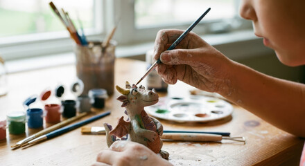 A boy paints a dinosaur toy at a table with art supplies in daylight.