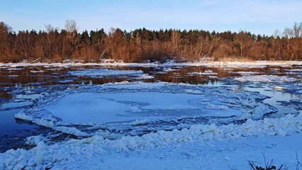Winter River with Drifting Ice Floes at Sunset. Serene winter river scene with numerous floating ice chunks on water surface reflecting warm sunset light against forested shore. © Anastasiya