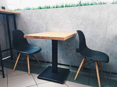 Arranging small wooden table with two black chairs against concrete wall indoors. Positioning compact dining setup featuring light wood tabletop and modern plastic chairs on tiled floor in minimalist 