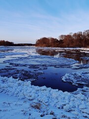 Winter River Landscape with Floating Ice Floes. Wide view of river partially covered with drifting ice chunks under clear blue sky with distant tree line. © Anastasiya
