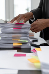 African American businesswoman organizing colorful paperwork stack on modern office desk with documents and laptop under soft daylight creating productive atmosphere for finance, consulting, HR
