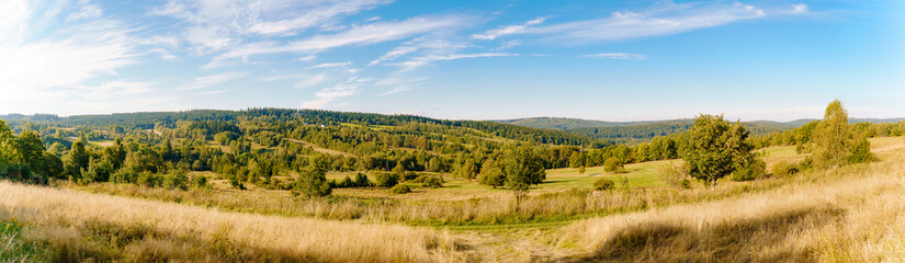Rural summer landscape panorama of the G&oacute;ry Sowie (Owl Mountains), Poland, featuring rolling green hills, open meadows, dense forests, scattered farms, country roads, and a clear blue sky.
