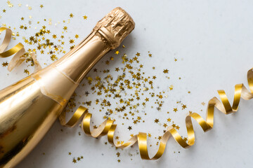 Gold champagne bottle and curling ribbon with star confetti on white marble. Flat lay party celebration background with copy space for text