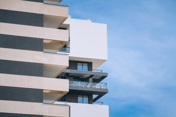 Modern apartment building with balconies and a clear blue sky. The structure features a mix of white and gray colors, showcasing contemporary architecture.