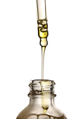 Studio shot of a glass pipette pouring a stream of golden cosmetic oil into an open glass bottle, isolated on a white background