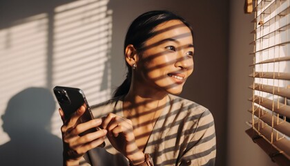 Happy young Asian woman holding a smartphone looks thoughtfully out a window, with beautiful warm sunlight creating striped shadows from the blinds across her smiling face