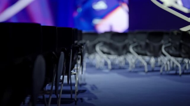 Empty conference auditorium with rows of modern chairs under blue and purple stage lighting, shot from a low aisle perspective as the venue awaits attendees for a presentation.