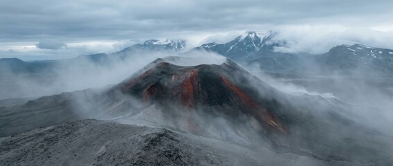 Steaming volcanic cone with red and black slopes is shrouded in thick mist against a backdrop of snow-capped mountains under a dramatic, cloudy sky