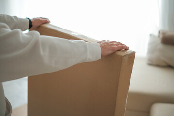 Woman hands holding large cardboard box with new furniture in bright, airy living room with sofa