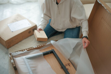 A young Asian woman unpacks a cardboard box at home. She is wearing a light-colored sweater and jeans. The scene shows a partially opened package and a wooden item.