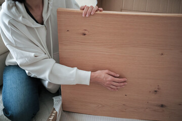 Mature woman in white hoodie examines new wooden table top. She is seated on floor and holding the board with both hands, showcasing its texture and grain.