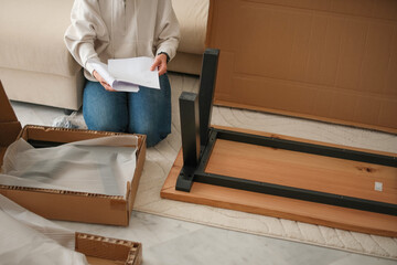 Mature woman sits on the floor, assembling furniture. She holds instructions and is surrounded by a cardboard box and wooden pieces of new wooden table