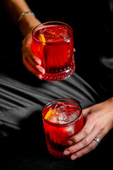 Two hands holding vibrant red Negroni cocktails in crystal rocks glasses with orange peels on a dark, elegant background