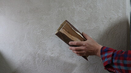 A man's hand is shown using a homemade device made of a small wooden board and sandpaper to clean and smooth plaster on a room wall; the DIY sanding tool is in the man's hand, scraping the plaster