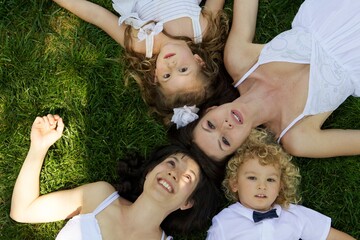 A happy family of four, including two adults and two children, lies playfully on lush green grass with their heads together, smiling up at the camera during a bright day.