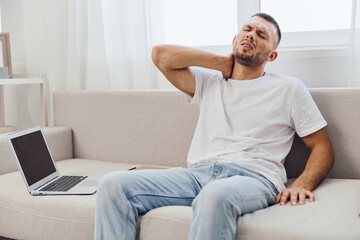 Tired man experiencing neck pain while working from home, wearing casual clothing, surrounded by a cozy, bright living space. This image captures the challenge of remote work.