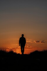 A powerful silhouette of a solitary individual walks purposefully towards the horizon, bathed in the warm, dramatic glow of a sunset or sunrise. The vibrant orange and deep amber sky, dotted with soft