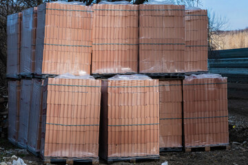 A static outdoor shot displays multiple stacked pallets of red clay building bricks wrapped in plastic film at a construction material supply yard.