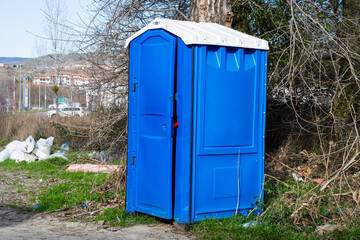 a blue portable toilet standing on a grassy patch near a tree, with trash scattered around it.
