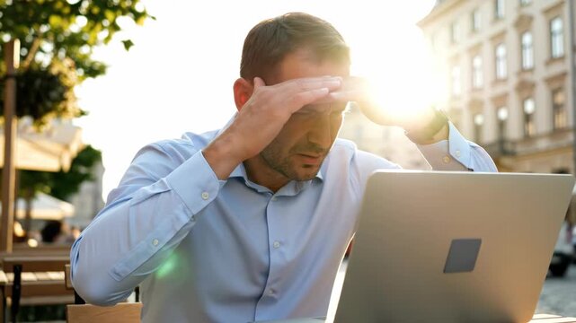 Man working on laptop at outdoor cafe. Bright sun glare causing frustration while typing. Remote work challenges during golden hour