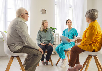 Group of elderly people sitting on chairs in circle with young female caregiver in bright room. Happy seniors sharing thoughts during therapy session, fostering emotional support and connection.