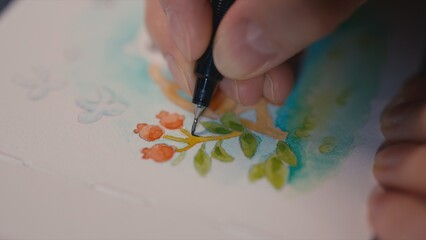 Close up of artist fingers outlining miniature red berries in whimsical watercolor painting with ink pen. Detail of park bench on green lawn, macro shot, selective focus on pen tip, blurred background