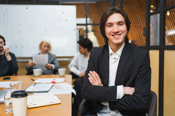 Fototapeta premium Portrait of team leader businessman in office, colleagues meeting in background. Confident business leader man sitting in office. Boss leader professional worker. Working process office life career