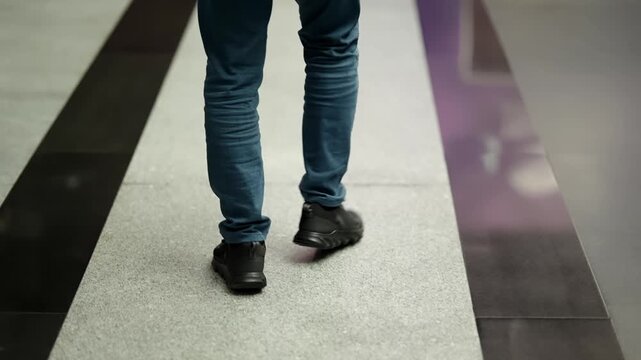 Close-up view of legs in blue jeans and black sneakers walking along a polished indoor corridor, highlighting motion of footsteps across contrasting gray and black floor panels