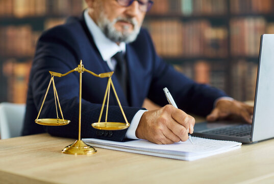 Lawyer writing notes in notebook, attorney working on clients defense in court case. Professional senior advocate in suit using laptop at desk with justice scales in law office, preparing for hearing