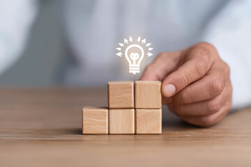 Wooden blocks stacked in step formation on table with hand placing top block, symbolizing growth and ideas with glowing light bulb icon above