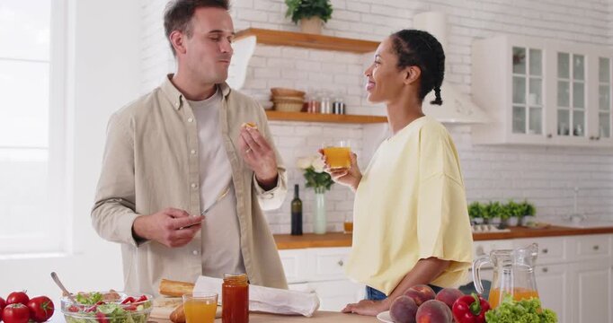 Couple cooking breakfast together in bright kitchen. They spread jam on bread, pour juice, and chat at home beside fruit and salad together. Cozy morning breakfast, love and togetherness.