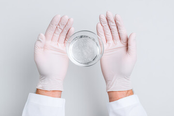 Clear liquid in glass bowl held by hands wearing pink gloves on white background, minimal style close up of hands mixing two clear skincare liquids with gentle care and precision