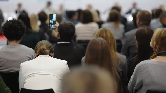 Seated audience at a professional conference, viewed from behind, as attendees focus on a distant panel discussion while some record on smartphones in a crowded seminar hall