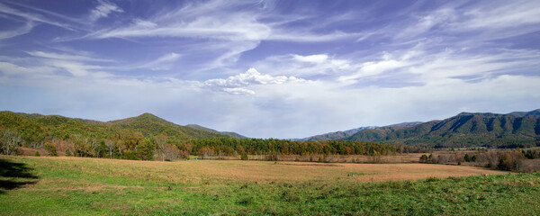 A couple sits on a blanket in Cades Cove, Tennessee. They set up a picnic under the sun with clouds in the sky. Green hills create a beautiful background around the meadow.