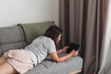Obraz premium Woman lying on sofa at home and browsing tablet screen while relaxing alone near window during quiet daytime leisure and casual indoor lifestyle concept