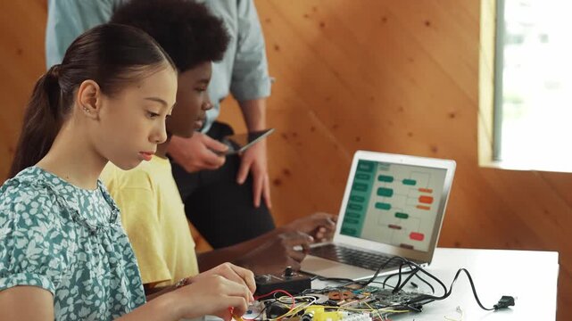 Teacher standing while explain coding and robotic construction. Diverse teenager fixing mother board and using electronic tool while looking at laptop display programming code screen. Edification