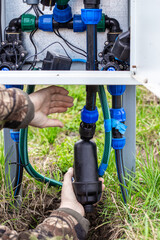 The hands of a person working on installing a water filter connected to a central system of pipes and fittings in a cabinet for automatic drip irrigation.