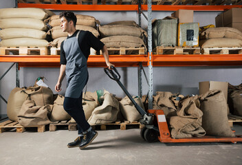 Male in apron pulling weighing pallet truck with coffee bags. Warehouse worker moving hydraulic...