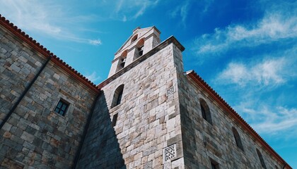 Fototapeta premium Historic stone church bell tower with terracotta roof tiles and visible bells, viewed from a low angle against a beautiful blue sky with wispy clouds on a sunny day