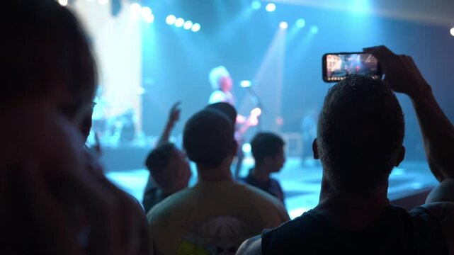Silhouetted audience members watch a live band on stage under bright blue spotlights. A raised smartphone records the concert in an indoor arena.