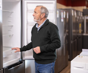 Fototapeta premium Elderly man buyer chooses electric refrigerator in home appliance store