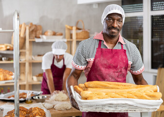 Smiling successful African American baker holding rustic wicker tray of golden crispy baguettes, proudly presenting freshly baked goods in cozy private bakery environment..