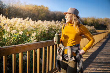 Woman standing on wooden boardwalk with reusable cup, autumn lake landscape, warm sweater and hat, quiet outdoor leisure, calm lifestyle concept