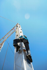 Adventurous person scaling tall metal mast structure against blue sky. High-altitude climbing activity showing courage and extreme recreation. © Victor