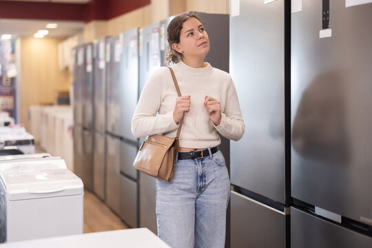Young woman stands in a hardware store and chooses a large fridge with freezer. Purchase of modern kitchen appliances