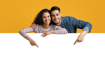 Happy Young Arab Couple Pointing Down At Big White Advertisement Board, Smiling Middle Eastern Spouses Showing Free Copy Space For Your Ad Or Design While Posing Over Yellow Studio Background © Prostock-studio