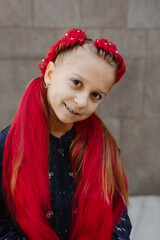 Young girl with elaborate red braided updo hairstyle with long synthetic hair extensions decorated with pearl beads, against gray tiled wall