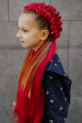 Young girl with elaborate red braided updo hairstyle with long synthetic hair extensions decorated with pearl beads, against gray tiled wall