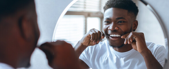 A man stands in front of a bathroom mirror, using dental floss to clean his teeth. Sunlight streams...